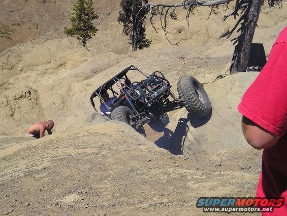 Off-road buggy with a roll cage climbing a steep, rocky hill. A person is partially submerged in the dirt to the left