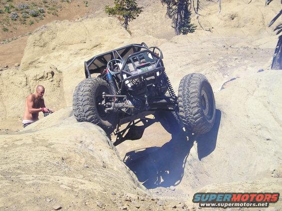 Off-road buggy with a roll cage and large tires navigating a rocky incline, a man stands nearby