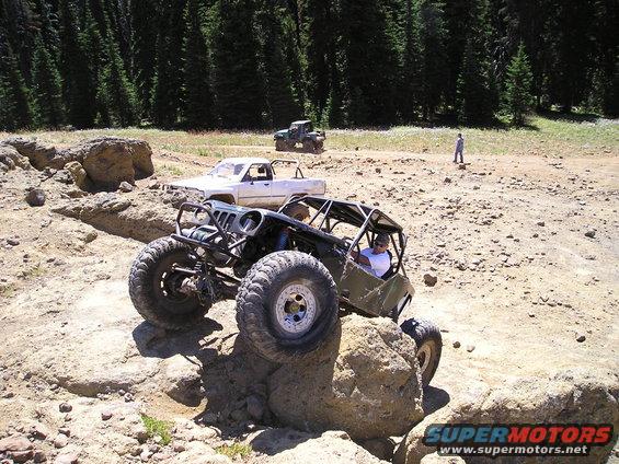Custom buggy with roll cage climbing over a rock, white pickup and green ATV behind it
