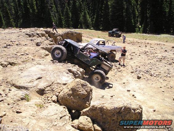 Off-road buggy climbing over rocks with a white pickup truck and another vehicle in the background