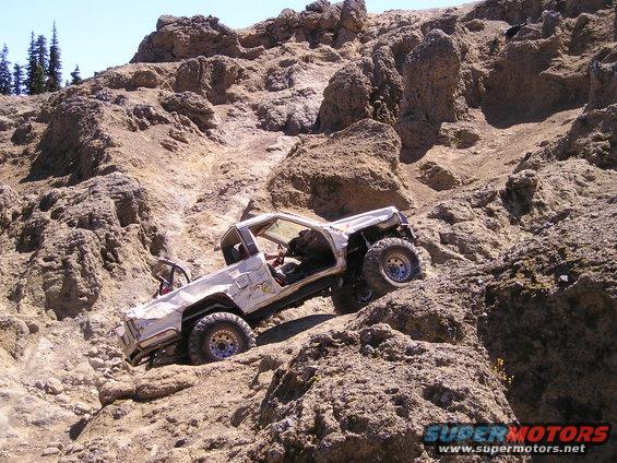 1989 Ford Bronco climbing a steep, rocky hill. The vehicle is white with off-road modifications
