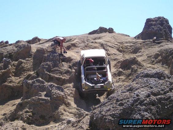 White SUV with roll cage climbing a steep, rocky hill. A person stands nearby. The vehicle's rear is visible