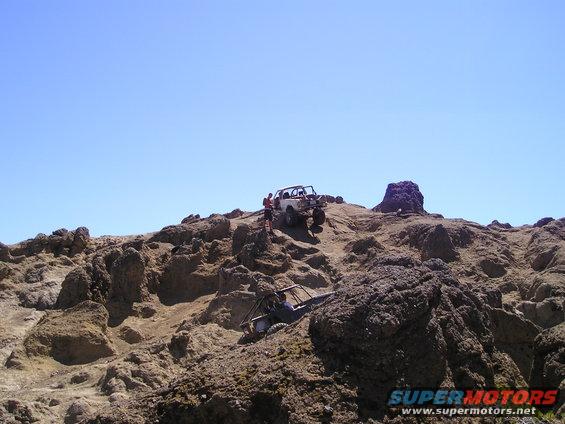 Two off-road vehicles navigating a steep, rocky hill under a clear blue sky