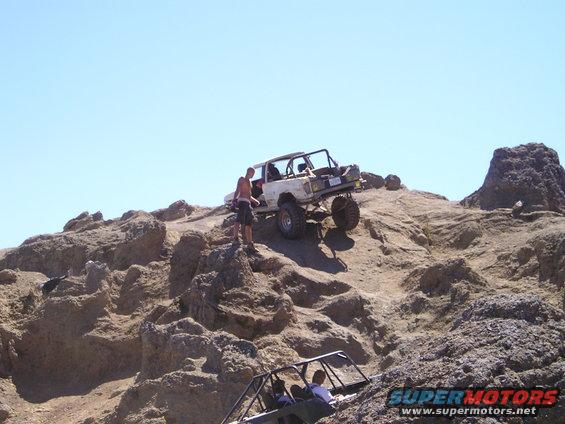 Two off-road vehicles are climbing a rocky hill. One white vehicle is higher up with a person standing next to it