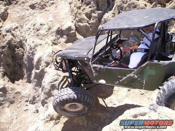 Off-road buggy with roll cage navigating rocky terrain. Driver wears a camo hat