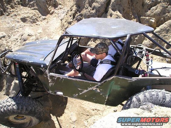 Off-road buggy with a driver and passenger navigating a rocky trail. The vehicle has a roll cage and large tires