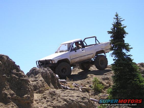 White pickup truck with large tires climbing a rocky hill. Driver is visible
