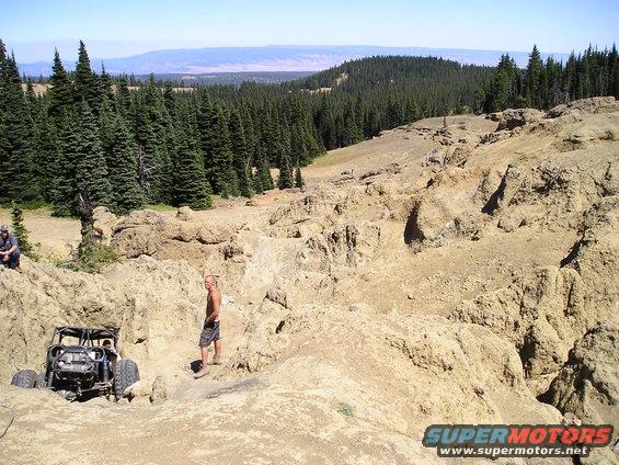 Off-road buggy navigating a rocky, uneven landscape with pine trees in the background. Two men are visible near the vehicle