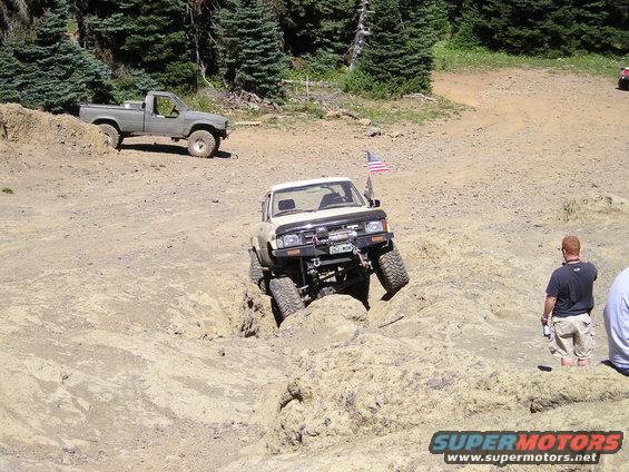 tan Toyota pickup truck navigating a rocky obstacle, gray pickup truck in background, people watching