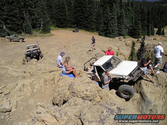 White off-road vehicle with open doors tilted on rocky terrain, people around