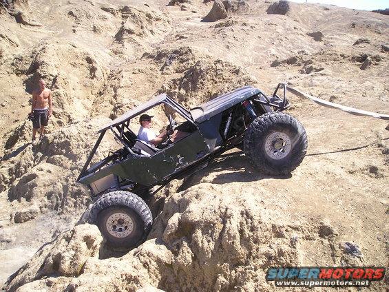 Custom buggy with roll cage and large tires navigating a rocky incline. Person stands on a nearby ledge
