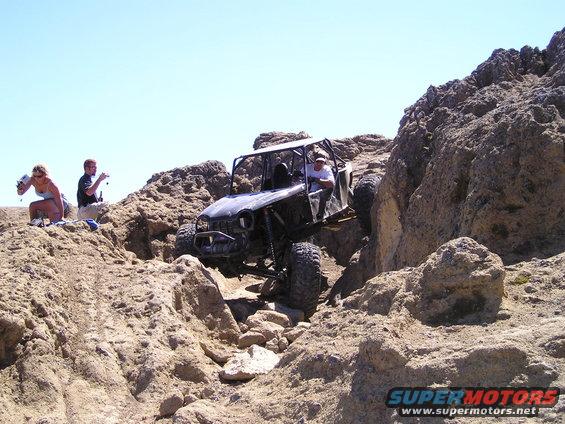Off-road buggy with roll cage climbing over rocks, driver visible