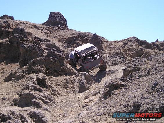 Gray off-road vehicle with a canvas top descends a steep, rocky slope under a clear blue sky