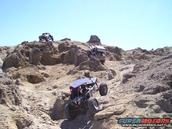 Three off-road vehicles navigating a steep, rocky hill under a clear blue sky. One vehicle is in the foreground, two are further up the incline