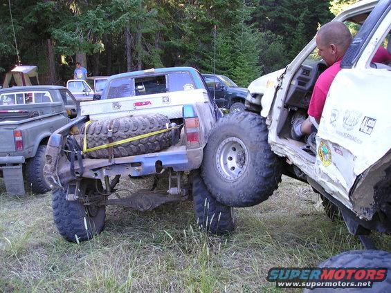 Two heavily modified trucks are stuck together off-road, one with a spare tire mounted on its rear