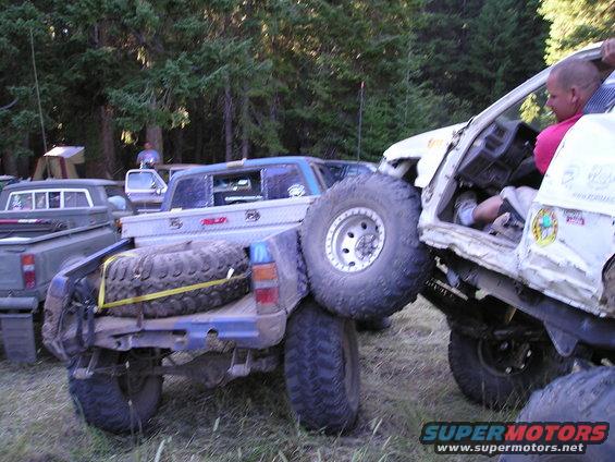 Two off-road trucks are stuck together, one on top of the other, in a wooded area. A spare tire is strapped to the bed of the lower truck
