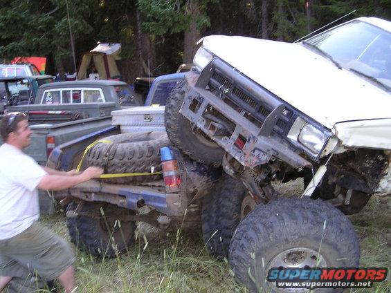 Two off-road trucks are stuck in grass, one with a man securing a spare tire to its bed
