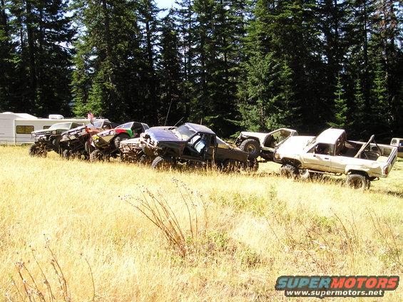 Several modified off-road trucks and SUVs parked in a dry grassy field with trees and a trailer in the background