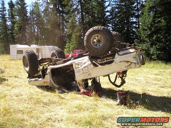 Rolled 1989 Ford Bronco on its roof, white, with large tires. Campers and trees in background