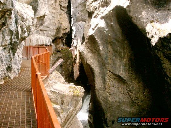 Metal walkway with orange railing alongside a narrow canyon with a small waterfall visible