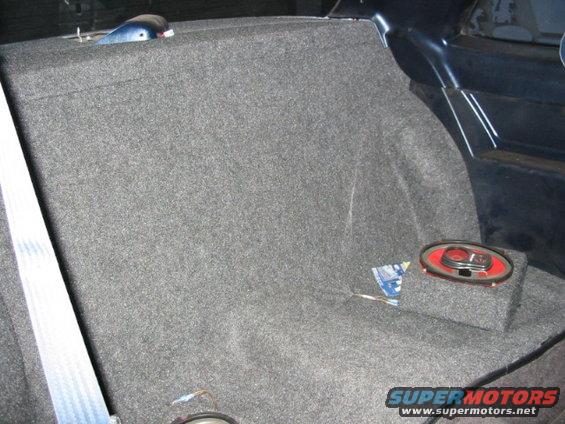 Car interior showing carpeted rear seat area with an aftermarket speaker box and oval speaker installed