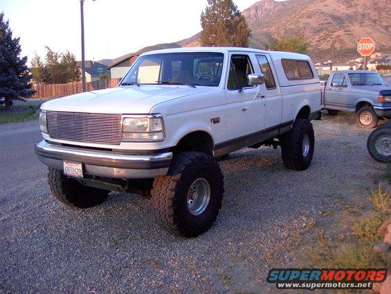 White 1992 Ford F-150 pickup truck with a camper shell, lifted with large tires, parked on gravel