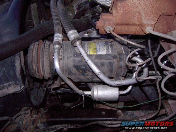 Close-up of a vehicle's AC compressor with hoses and a filter drier