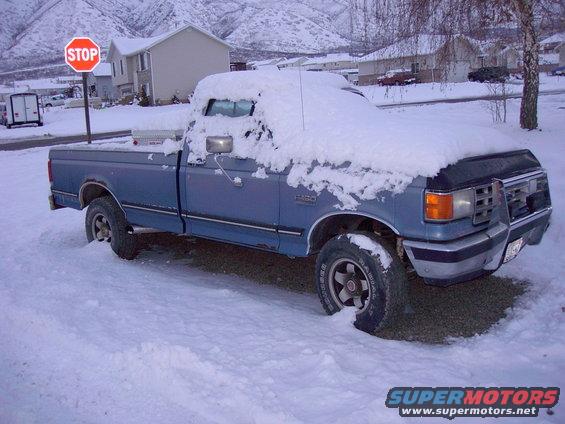 Blue 1992 Ford F-150 pickup truck covered in snow, parked on a street with houses and mountains in the background