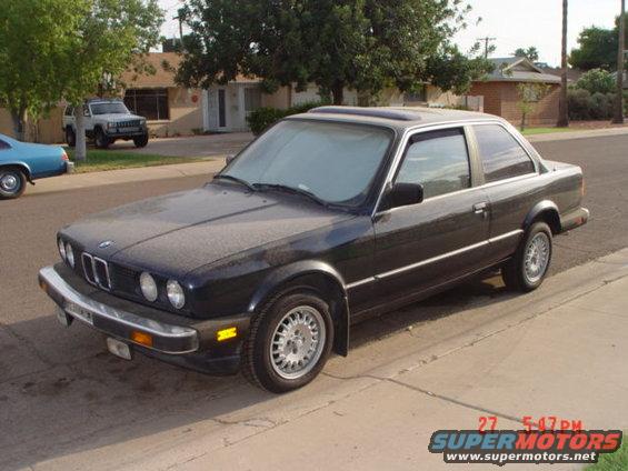 Dark BMW E30 coupe parked on street, white SUV in background