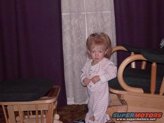 Toddler in striped pajamas stands near a wooden rocking chair and a cushioned bench