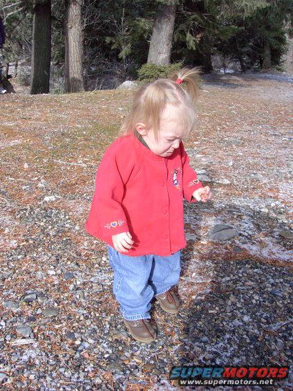 Toddler girl in red jacket and jeans standing outside on a rocky, wooded area