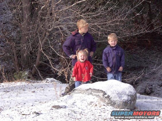 Three children stand in a snowy wooded area. One child in red is crying. A large rock is in the foreground