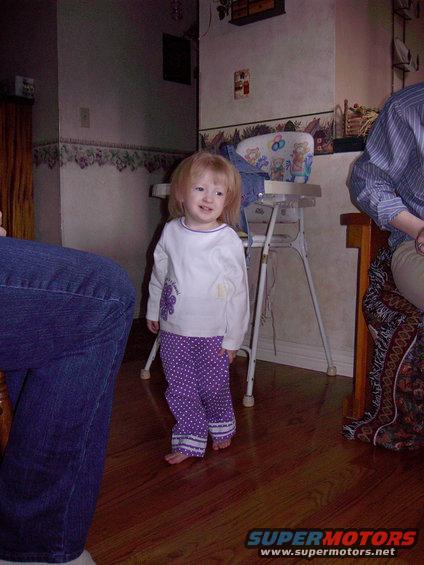 Toddler girl in purple polka dot pants and white shirt stands in a room with wooden floors