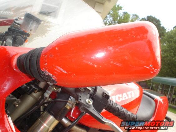 Red motorcycle mirror with black bellows and scuff marks, close-up on handlebars and part of the 'DUCATI' logo