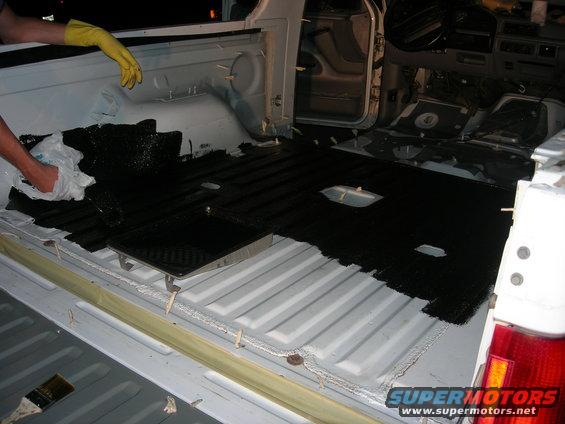 Truck bed interior with white ribbed walls, being coated with black material. A person in yellow gloves is visible