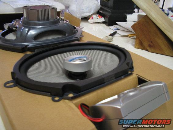 Two car speakers and a silver crossover with red wires sit on cardboard