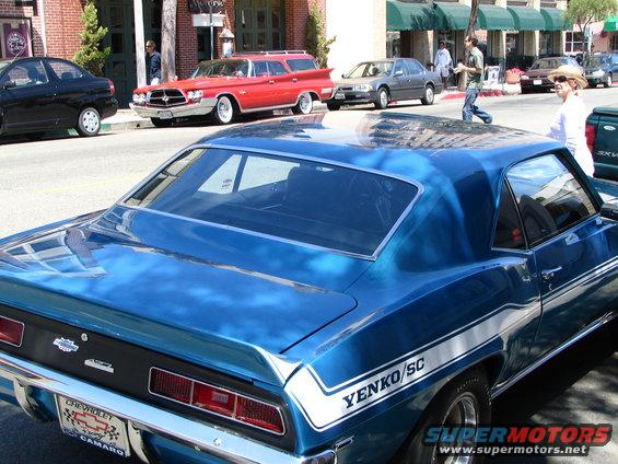 Blue 1969 Chevrolet Camaro Yenko/SC with white stripe, seen from rear. Other cars and buildings in background