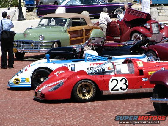 Red race car with number 23 and white stripe, next to a white and blue race car with number 11. Other classic cars are in the background