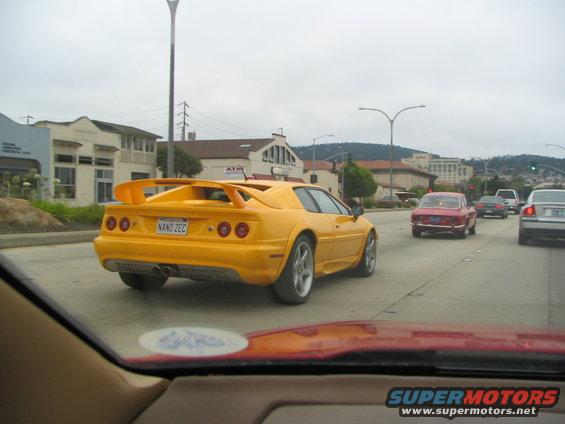 Yellow sports car with a big wing and a license plate that says NANO ZEC, driving on a cloudy day