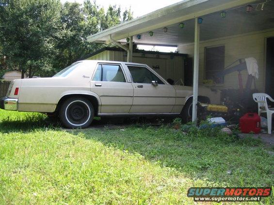 tan sedan parked under a carport, overgrown grass in front