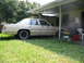 1985 Ford Crown Victoria LTD - A tan sedan is parked under a carport with overgrown grass in the foreground.