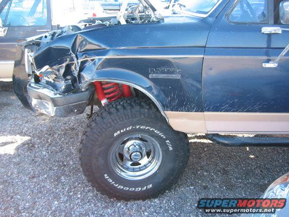 Front end of a blue 1989 Ford Bronco with significant damage. Red shocks are visible