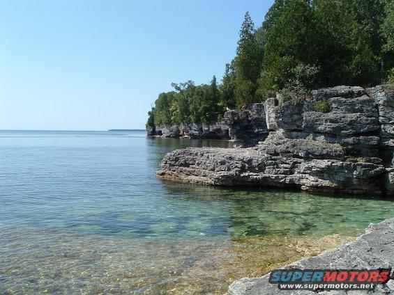 Rocky coastline with clear turquoise water and trees under a blue sky. Watermark reads SUPERMOTORS