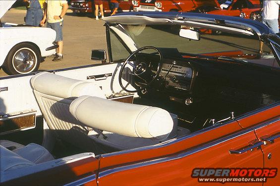 Interior of a red convertible with white seats, showing the dashboard and steering wheel. Other cars are visible in the background