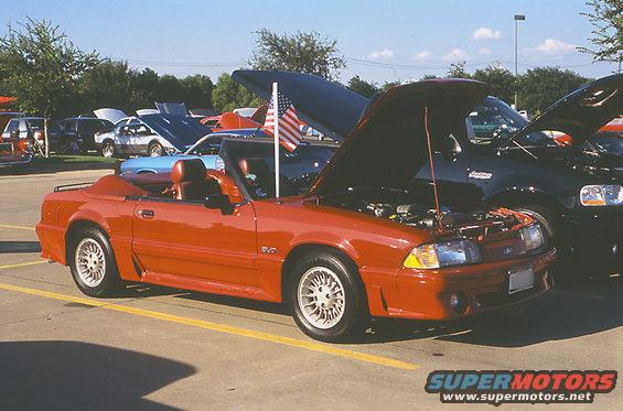 Red convertible Mustang with hood open, parked at an outdoor car show. American flag on a pole is visible