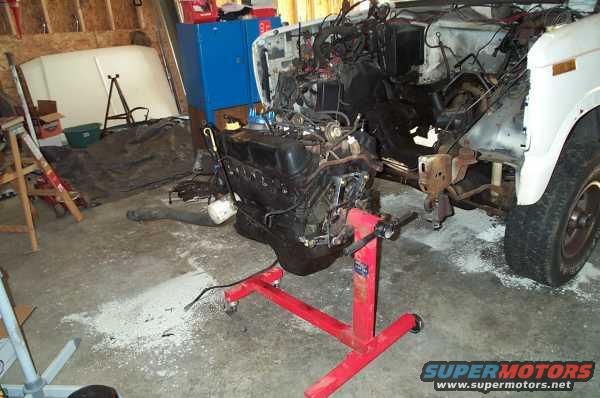 Engine on red stand in garage, next to a white vehicle with its hood removed and engine bay exposed