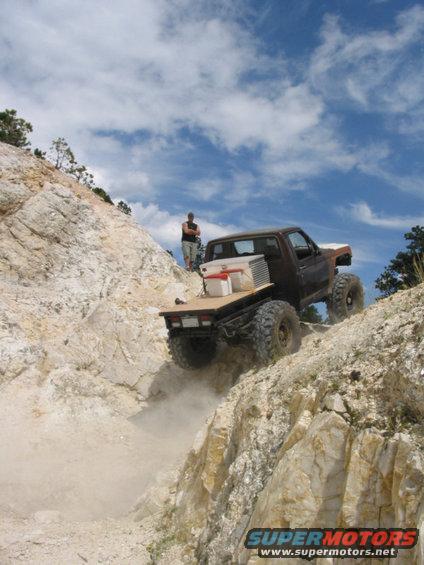 Truck climbing a steep, rocky hill with a flatbed in the back. Person standing on the hill watching