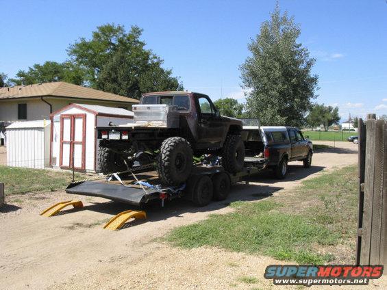 Brown off-road truck with large tires on a flatbed trailer, towed by a dark pickup truck