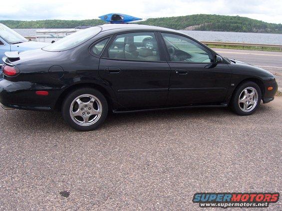 Black sedan parked by a lake, with a blue kayak on its roof. Another car is partially visible