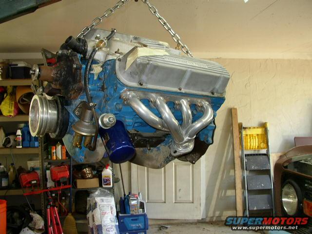 V8 engine with blue block and aluminum valve covers hanging from chains in a garage